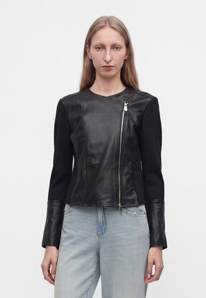 Femme aux longs cheveux blonds portant une veste en cuir noire avec une fermeture éclair asymétrique et un jean bleu clair sur fond uni.
