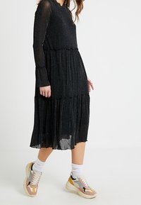 Svart långärmad klänning med en glittrande yta, volangkjol och elastiska ärmslut, i kombination med flerfärgade sneakers och vita ankelsockor.