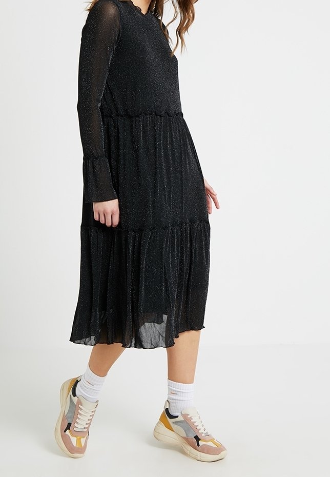 Svart långärmad klänning med en glittrande yta, volangkjol och elastiska ärmslut, i kombination med flerfärgade sneakers och vita ankelsockor.
