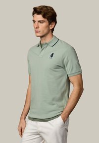 Mladý muž v svetlozelenej polo košeli s výšivkou čísla 4, bielych nohaviciach, stojaci s jednou rukou v vrecku na jednoduchom pozadí.