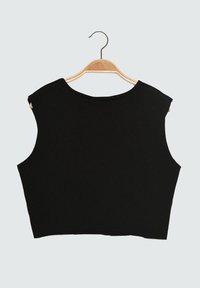 Top court sans manches noir avec de larges emmanchures suspendu sur un cintre en bois contre un fond léger uni.