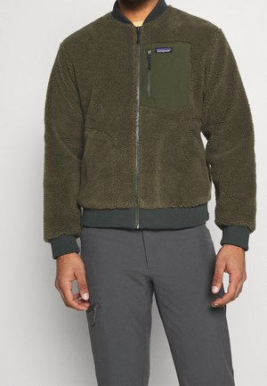 Mężczyzna ubrany w oliwkowo-zieloną polarową bluzę na zamek z kieszenią na piersi i ciemnoszare spodnie, stojący na tle jednolitego jasnego tła.