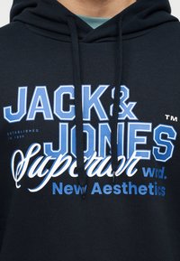 Κοντινό πλάνο ενός ατόμου που φοράει μαύρο φούτερ με κουκούλα, με μπλε και λευκό κείμενο "JACK & JONES Support New Aesthetics" στο στήθος.