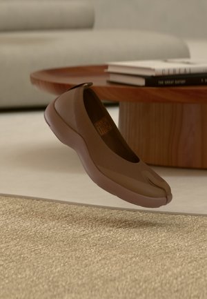 Chaussure marron à enfiler avec un design à bout fendu flottant au-dessus d'un tapis beige tissé, à côté d'une table ronde basse en bois avec des livres empilés.