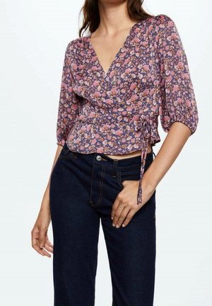Woman wearing a purple floral wrap blouse with puff sleeves and dark blue jeans, her right hand resting on her hip.