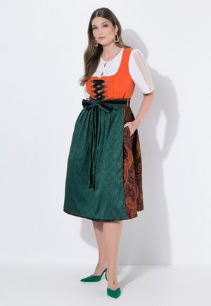 Donna che indossa un vestito tradizionale con corpetto arancione, camicetta bianca, grembiule verde legato con un fiocco e tacchi verdi, in piedi con la mano in tasca.