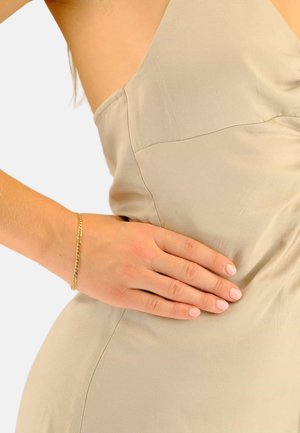 Robe beige avec un corsage drapé, en tissu lisse. Le poignet arbore un bracelet délicat en chaîne d'or. La peau est visible sur le côté.