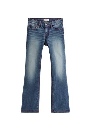 Teenager står frontalt, iført en tætsiddende hvid T-shirt med korte ærmer, blå udsvingede jeans og hvide sneakers i et simpelt studie.
