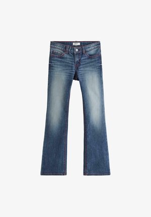 Teenager står frontalt, iført en tætsiddende hvid T-shirt med korte ærmer, blå udsvingede jeans og hvide sneakers i et simpelt studie.