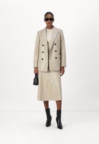Beige Blazer aus Kunstleder mit doppelter Knopfleiste in Schwarz, passendem Midirock, schwarzen Stiefeletten und dunklen Sonnenbrillen.