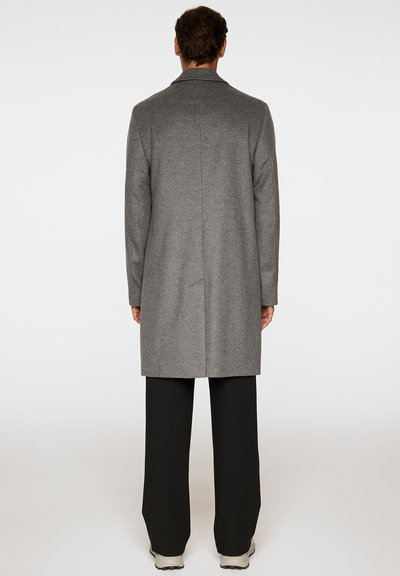 Uomo in piedi, girato di schiena, che indossa un lungo cappotto grigio, pantaloni neri e scarpe bianche, su uno sfondo leggero e neutro.