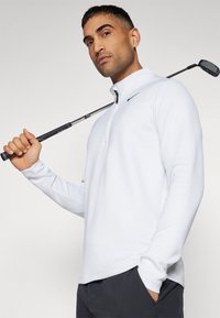 Pulover de golf alb, texturat, cu mânecă lungă, guler cu fermoar în patru sferturi și un logo negru. Modelul ține o crosă de golf peste umăr.