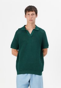 Polo shirt en maille verte à manches courtes, avec un col en V, un col côtelé et un motif texturé. Associé à un pantalon bleu clair.
