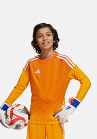 Oranje shirt met lange mouwen met een gestructureerd patroon, wit adidas-logo en drie witte strepen. Het kind houdt een voetbal vast in keepershandschoenen.