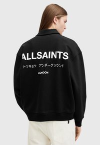 Musta ylipitkä huppari, jossa on korkea kaulus ja valkoista painettua tekstiä selässä: "ALLSAINTS," japaninkielistä tekstiä ja "LONDON."