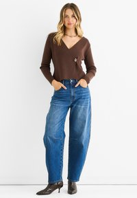 Ruskea neulottu kietaisupaita, jossa on V-pääntie ja koristeellinen nappi, yhdistettynä korkeavyötäröisiin sinisiin farkkuliiveihin ja kärjellisiin käärmeennahkakorkon kenkiin.