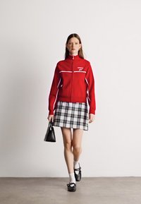 Chaqueta roja con cremallera y acentos blancos combinada con una falda mini de cuadros. El modelo lleva un pequeño bolso negro y usa calcetines blancos con zapatos negros.