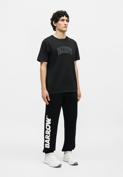 Νέος άνδρας με κατσαρά μαλλιά, φοράει μαύρο t-shirt Barrow και μαύρη φόρμα Barrow με λευκά sneakers, στέκεται μπρος σε απλό φόντο.