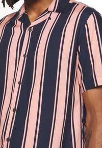 Camisa de manga corta, tipo boton-up, en rayas navy y rosa claro, fabricada con un tejido suave. Presenta un cuello de campamento y botones negros.