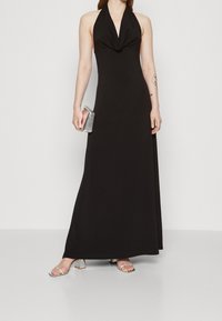 Black halter-neck maxi dress with a draped front, made from a smooth fabric. Paired with clear heeled sandals and a silver clutch.