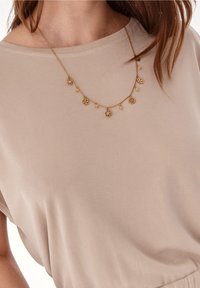 Bežový top s krátkým rukávem a kulatým výstřihem, který má jemný zlatý řetízek zdobený kulatými květinovými přívěsky a malými korálky.