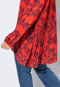 Camicia rossa a pieghe con motivi testuali neri, maniche lunghe con polsini elasticizzati, abbinata a pantaloni jeans blu, mostrando uno stile casual.