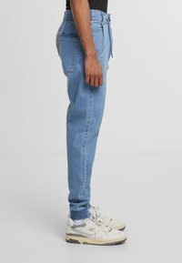 Svijetloplavi denim joggeri s suženim nogavicama, elastičnim pojasom i bočnim džepovima, uparenih s bijelim i sivim atletskim tenisicama.