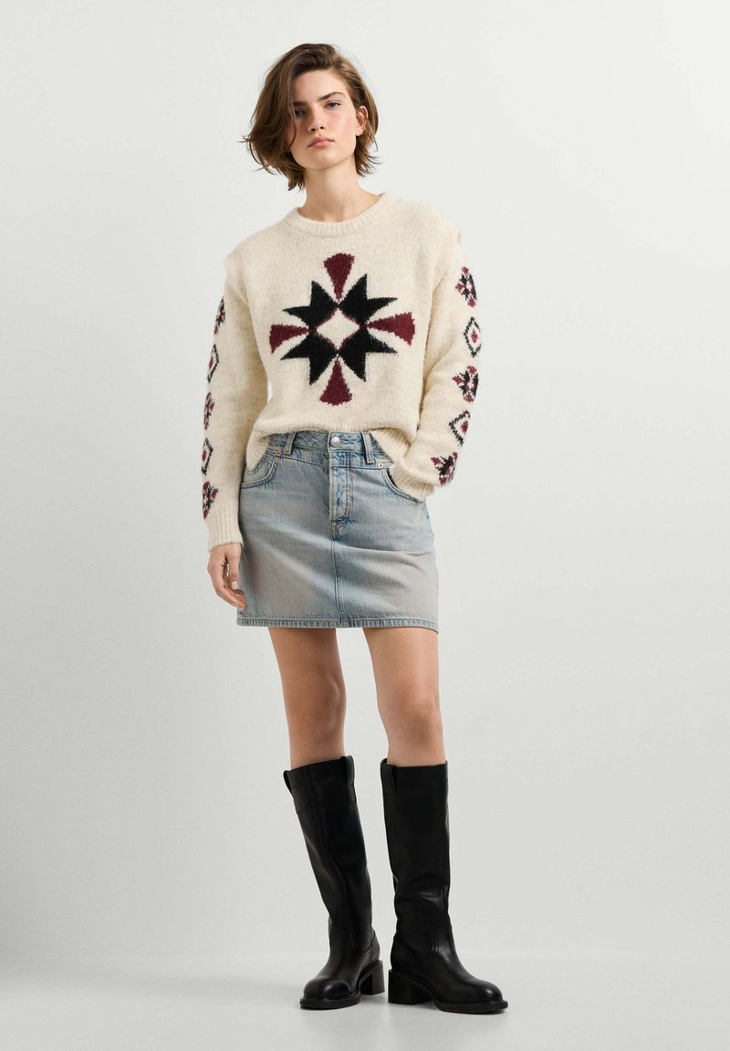 Femme portant un pull crème à motifs géométriques, une jupe en jean bleu clair et des bottes noires montant jusqu'aux genoux, debout devant un fond uni.