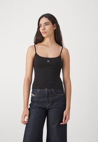 Jeune femme aux longs cheveux foncés portant un top noir à fines bretelles et un jean large bleu foncé sur un fond uni.