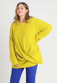 Sennepsgul, ribbet oversized genser med rund hals og vridd detalj foran, parret med blå bukser. Myk tekstur.