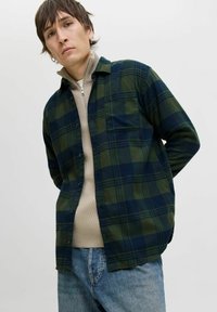 Junger Mann in einem grün-blauen Karoshemd über einem beigen Reißverschlusspullover und blauen Jeans, der vor einem schlichten hellen Hintergrund steht.