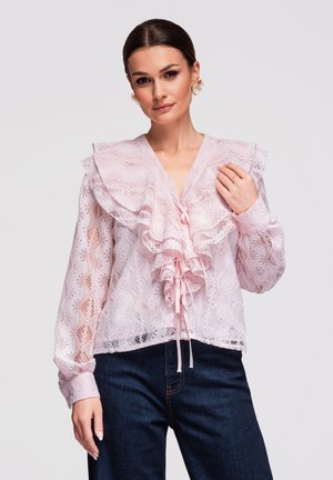 Femme portant un chemisier en dentelle rose transparent avec des volants superposés et des nœuds, associé à un jean bleu foncé sur un fond uni.