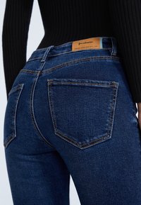 Dark blue skinny jeans with contrasting orange stitching, five pockets, and branded tan leather patch on the back waistband.