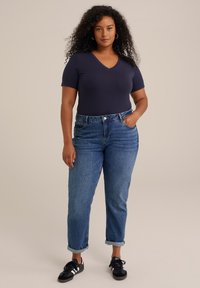 Camiseta de punto azul marino de manga corta y escote en V combinada con jeans de tiro alto de lavado medio. El calzado presenta zapatillas negras con rayas blancas.