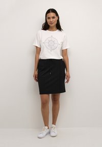 T-shirt blanc avec un graphique de compas, associé à une jupe noire avec une taille élastique et un cordon de serrage, portée avec des baskets blanches.