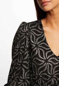 Svart mönstrad klänning med silverfärgade virvlande mönster, v-ringad hals och puffärmar. Strukturerad tyg ger djup åt designen.