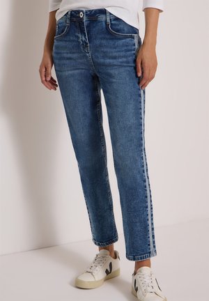 Vrouw draagt blauwe spijkerbroek met lichtere zijstrepen, witte sneakers met zwarte details en een wit shirt met lange mouwen.