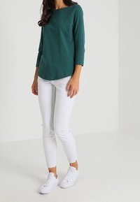 Vrouw in een donkergroene top met driekwartmouwen, witte skinny jeans met rafelige zomen en witte sneakers, staand tegen een effen achtergrond.
