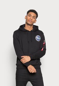 Fato preto com um bolso frontal, capuz com cordão e um emblema bordado em forma circular com um logótipo de foguete. Detalhe com zipper na manga esquerda.