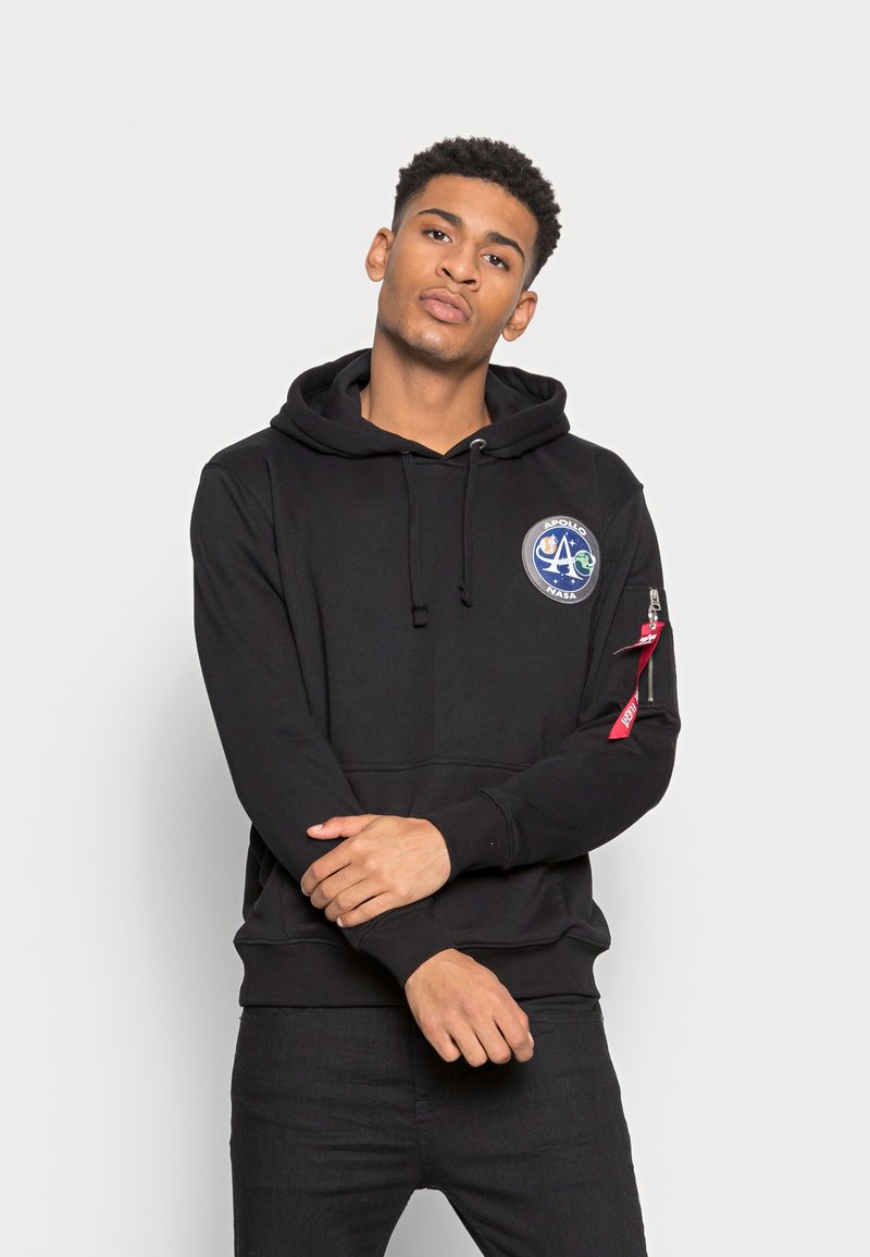 Fato preto com um bolso frontal, capuz com cordão e um emblema bordado em forma circular com um logótipo de foguete. Detalhe com zipper na manga esquerda.
