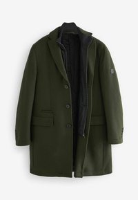 Manteau en laine vert foncé pour homme avec trois boutons, doublure intérieure zippée, revers crantés, poches à rabat et un écusson sur la manche gauche.