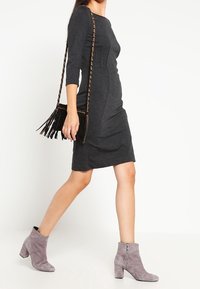 Šedé přiléhavé šaty s tříčtvrtečními rukávy, texturovaná látka, v kombinaci se šedými kotníkovými botami a kabelkou na rameno s třásněmi.