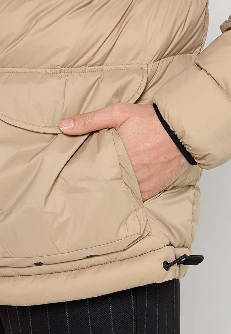 Beige puffjacka med glänsande finish, som har en sidficka, elastisk dragsnodd i fållen och en texturerad detalj på ärmslutet.