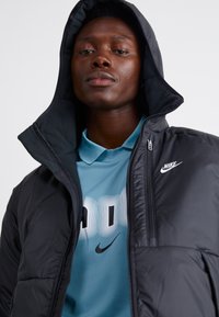Czarna pikowana kurtka z kapturem, z zamkiem błyskawicznym i logo Nike. Pod nią turkusowa koszulka polo z graficznym nadrukiem.