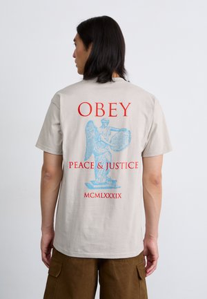 Beige t-skjorte med blå engelgrafikk og rød tekst "OBEY" og "PEACE & JUSTICE" med datoen "MCMLXXXIX" under. Korte ermer, avslappet passform.