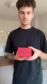 Sarkans taisnstūra kaste ar matētu apdari, uz kuras ir rakstīts "HUGO" treknrakstā pelēkā krāsā. To tur cilvēks pret neitrālu iekšējo fona.