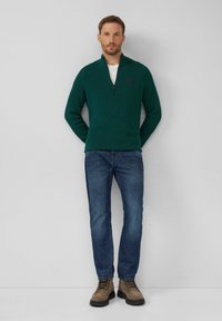 Dunkelgrüner strukturierter Pullover mit einem Reißverschlusskragen, kombiniert mit blauen Jeans und hellbraunen Schnürboots, mit einem Logo auf der Brust.