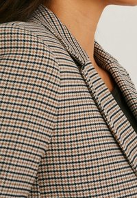 Teksturoidu bleiseri, jossa on musta, beige ja ruskea houndstooth-kuvio, notched-kaulus ja muotoillut olkapäät.