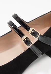 Černé semišové baleríny se dvěma lesklými strapcemi z černého lakovaného materiálu se zlatými sponkami, zdůrazňující minimalistický design a hladkou texturu.