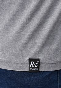 Šedé bavlněné tričko s hladkou texturou, s černou nášivkou na spodní části s nápisem "R:C RE.COVERED" v bílé barvě.